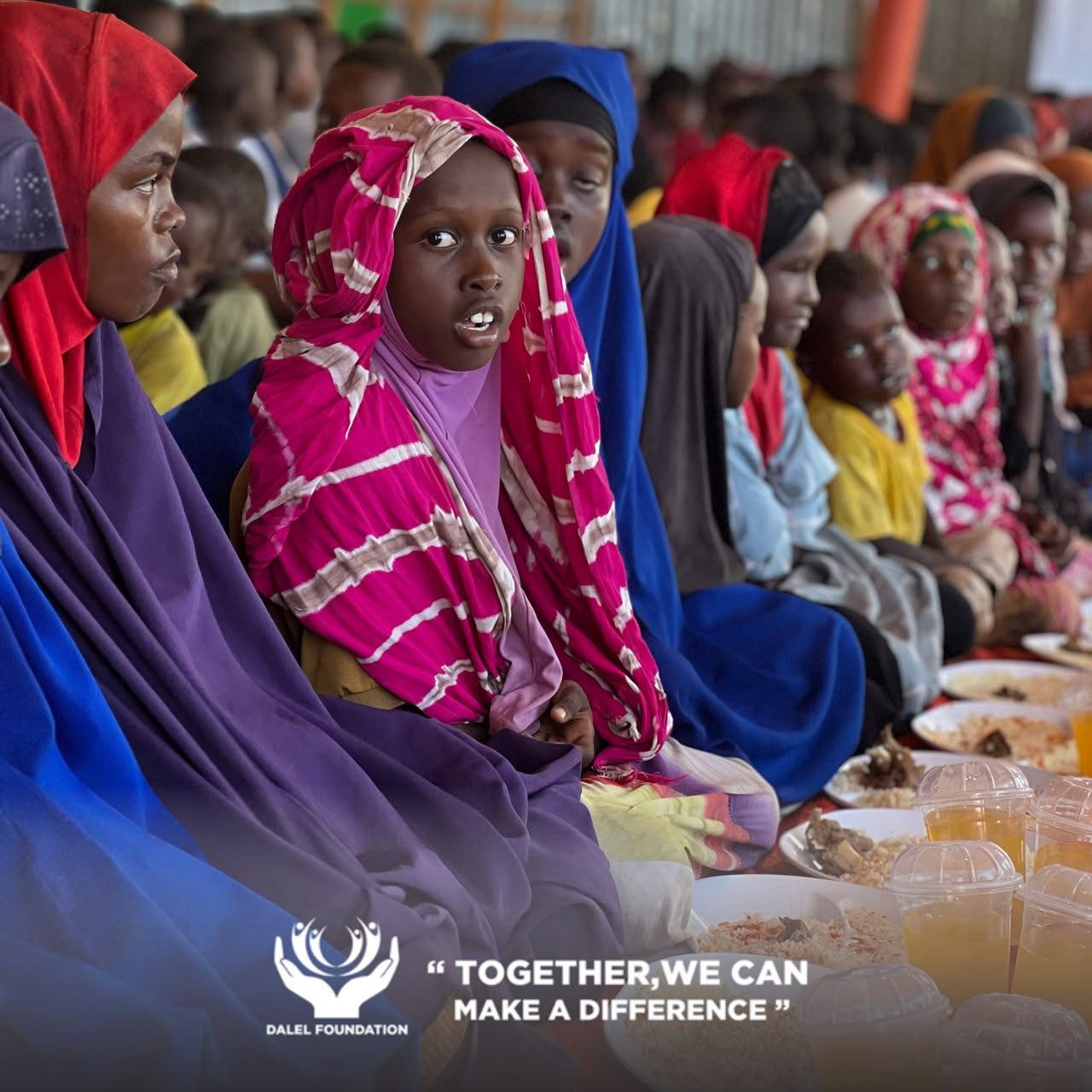 Women and children seated in a row beside plates of rice and fresh drinks at a Dalel Foundation meal distribution